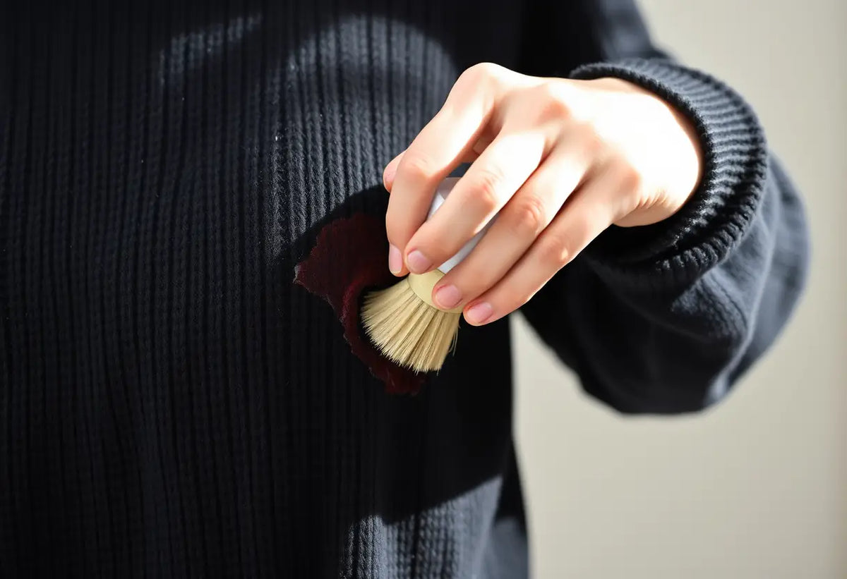 Une main applique une solution sur une tache d'un pull en laine noire, mettant en valeur le soin méticuleux nécessaire.
