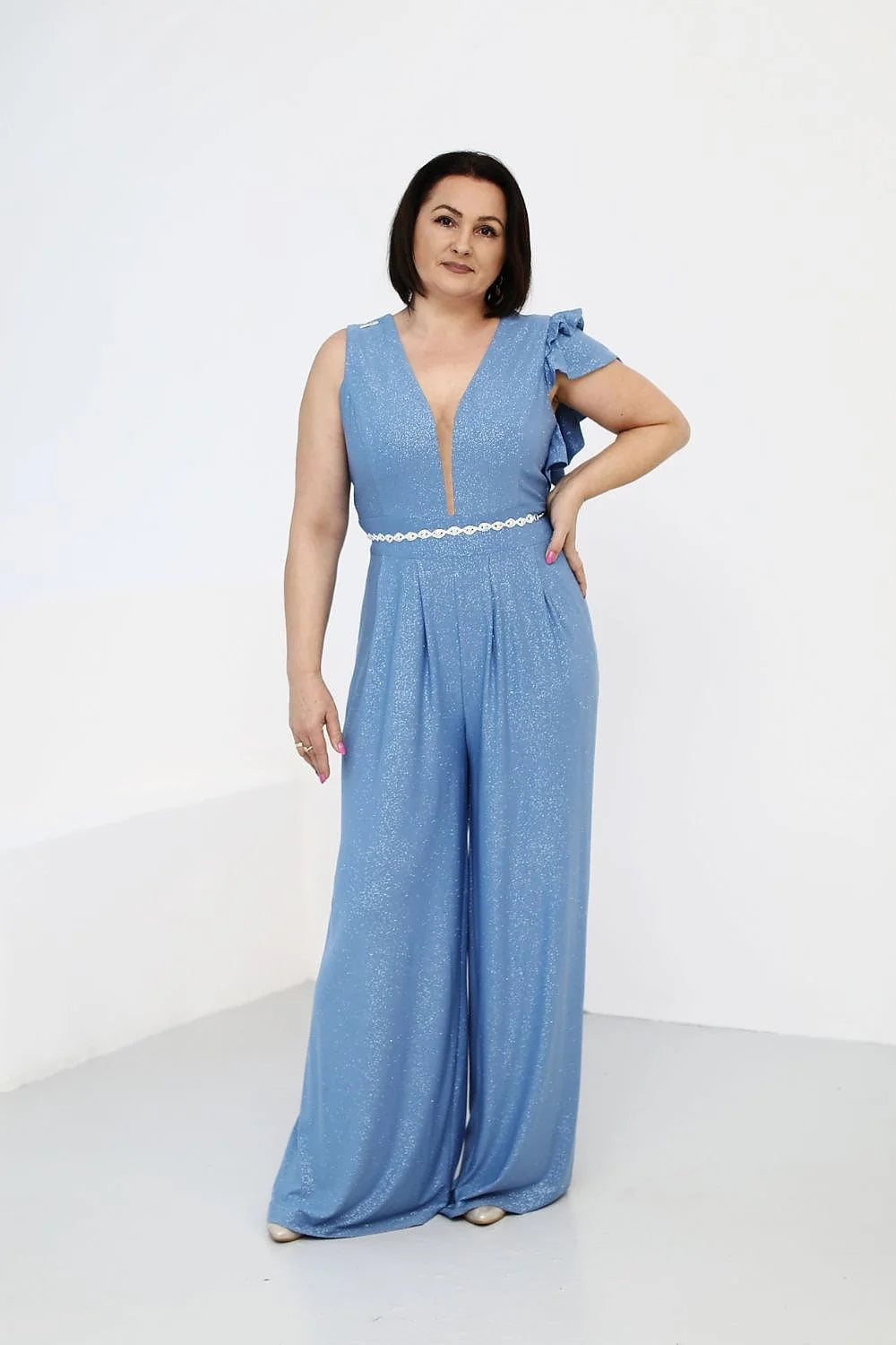 Combinaison élégante pour femme en tissu bleu clair scintillant avec encolure en V, manche asymétrique à volants, ceinture en dentelle blanche et jambes larges fluides